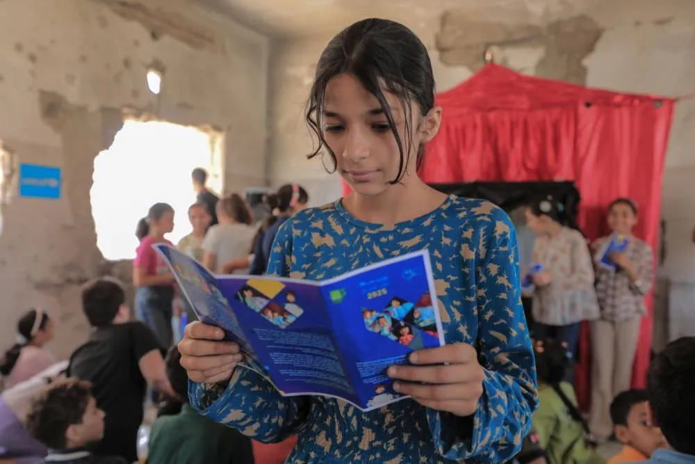 A girl reads a pamphlet in a room filled with other children.
