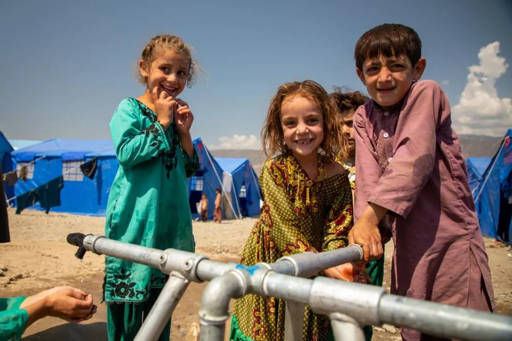 Children gathered around an outdoor water tap smile at the camera.