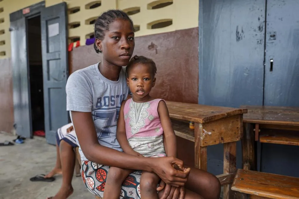 A person holding a child sits by a school desk and looks at the camera.