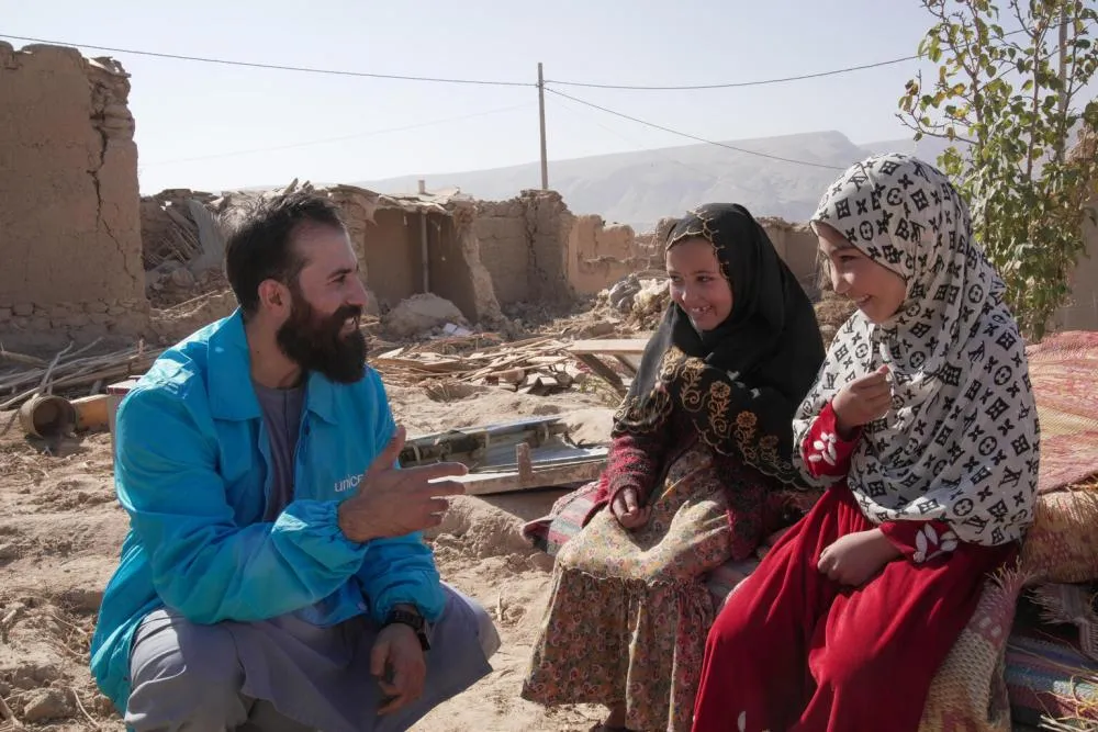 Outside amid rubble, two children wearing headscarves speak to a man wearing a blue UNICEF jacket.