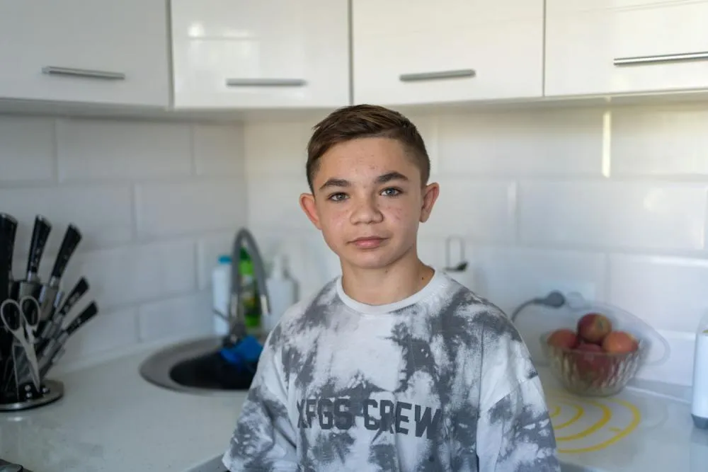 A boy standing in a kitchen looks at the camera with a neutral expression.