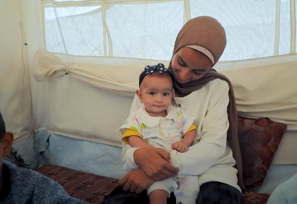 A mother is seated and holding an infant on their lap inside a tented shelter