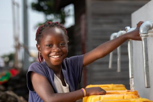 Une jeune fille sourit à l’objectif en puisant de l’eau à une fontaine.