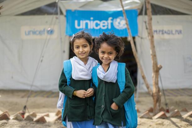 Two young girls wearing blue UNICEF backpacks smile at the camera while standing in front of a UNICEF-funded Temporary Learning Centre.