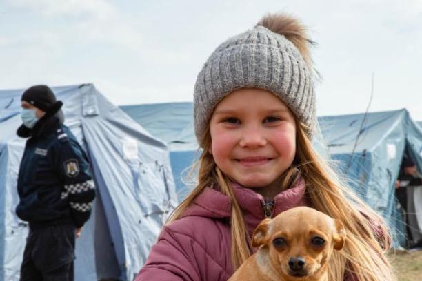 A young girl carrying a dog smiles at the camera in front of tents at a Temporary Refugee Centre on the border of Moldova and Ukraine.