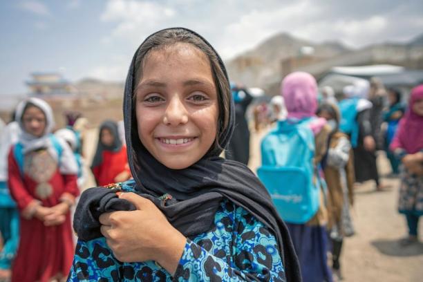In Afghanistan, a girl stands smiling at the camera, outdoors with her classmates in the schoolyard behind her wearing blue UNICEF backpacks.