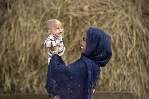 An infant laughs as he is playfully held up by his mother who is smiling back at him.