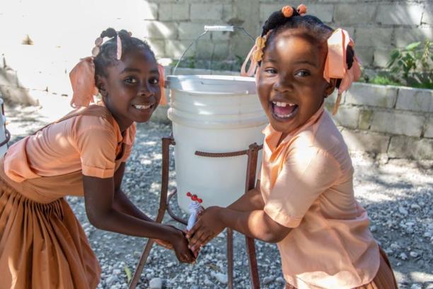 Two children in school uniforms smiling and looking at the camera while holding their hands under the spout of a water bucket.