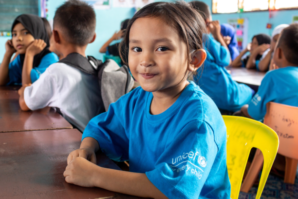 Portrait of a smiling child seated at a classroom table with their hands folded in front of them.