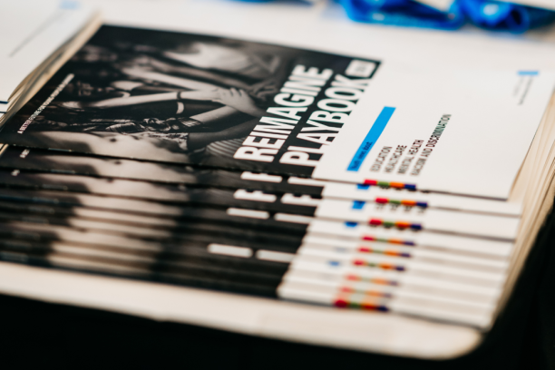 Closeup of report copies spread out on a table, with the title “Reimagine Playbook”.