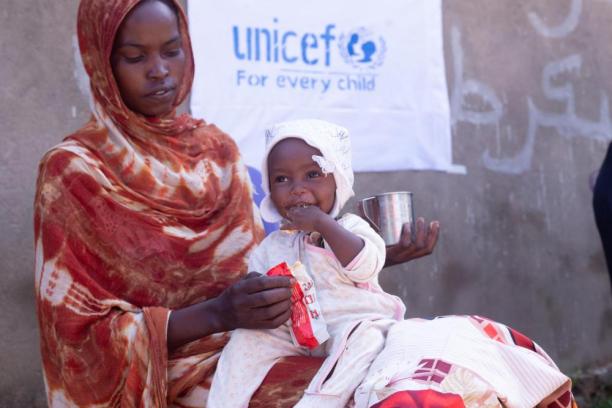 A woman holds a packet of therapeutic food while a baby sits on her lap. Above them is the UNICEF logo. 