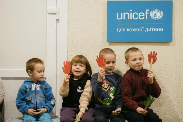 Children sit under the UNICEF logo and smile while holding crafts.