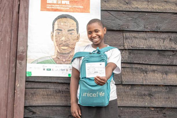 A girl with a UNICEF backpack holds up her vaccination card.