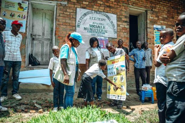 A group of people, including children and health workers wearing vests, gather around a large Ebola awareness poster showing symptoms and prevention measures.