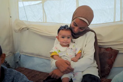 A mother is seated and holding an infant on their lap inside a tented shelter