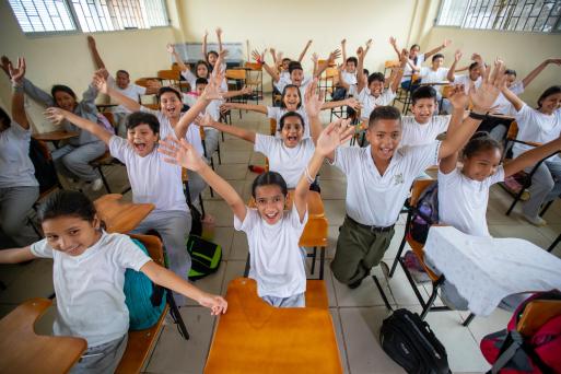 In a classroom, students smile and raise their arms. 
