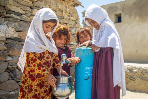 A group of girls stand around an outdoor water tap, one of them fills a jug with water.