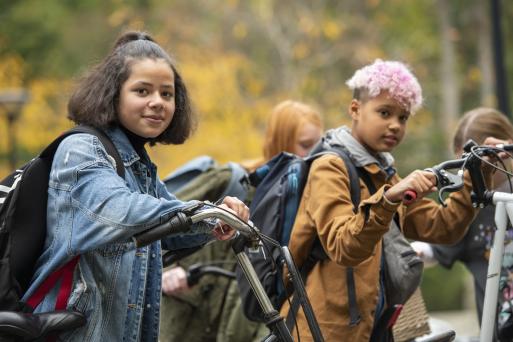 Deux enfants, une fille et un garçon, avec leurs bicyclettes, portant un sac à dos sur les épaules. Des arbres au feuillage jaune se dessinent à l’arrière-plan.
