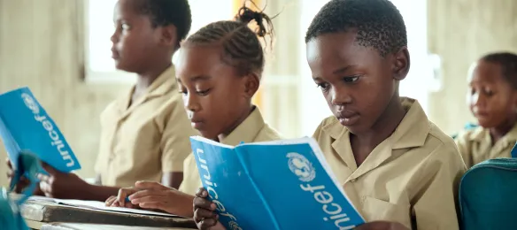 Children in school uniforms reading UNICEF exercise books at classroom desks.