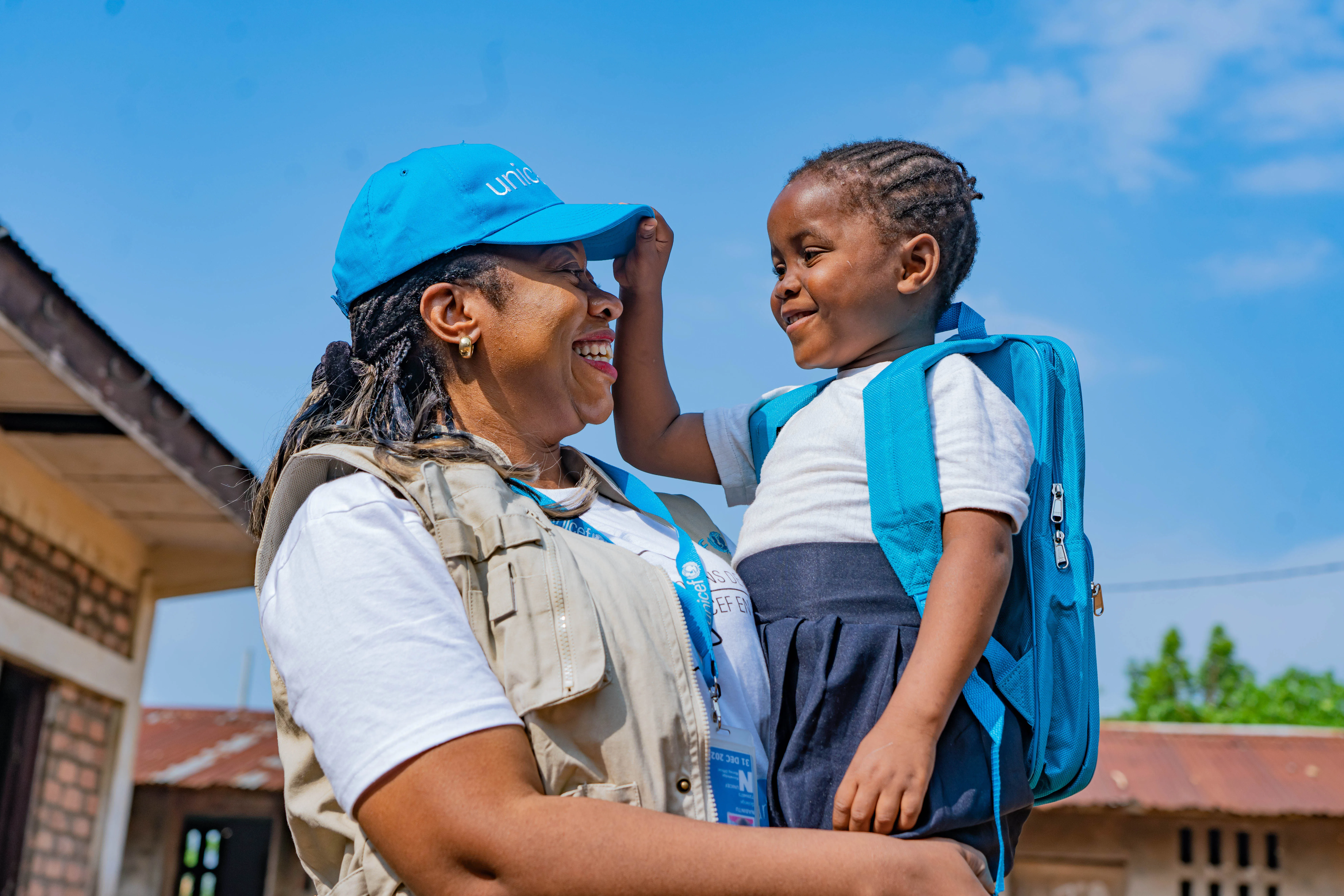 A smiling UNICEF worker stands outside in the sunshine holding a child who is smiling and reaching for the worker’s UNICEF hat.