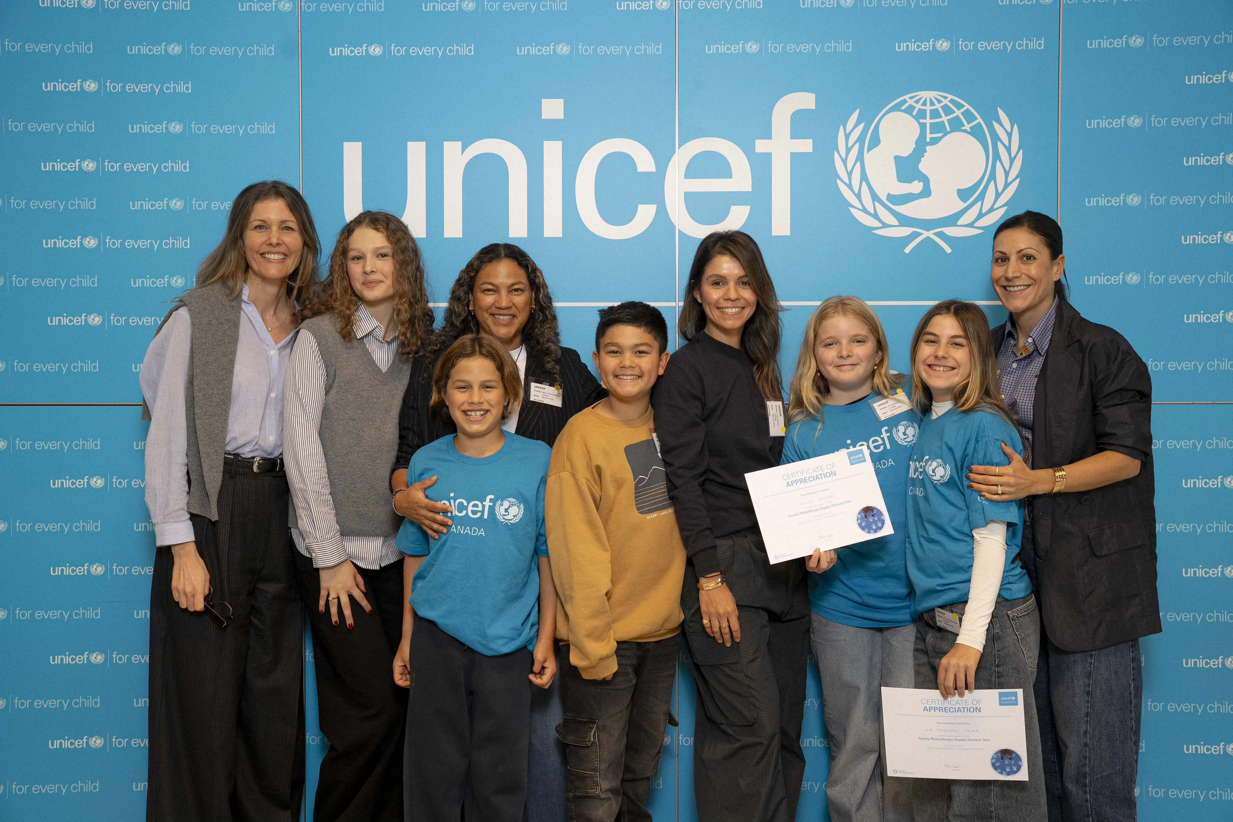 A group of four women and five children smile against a blue UNICEF backdrop. Two kids hold certificates. The scene is joyful and celebratory.