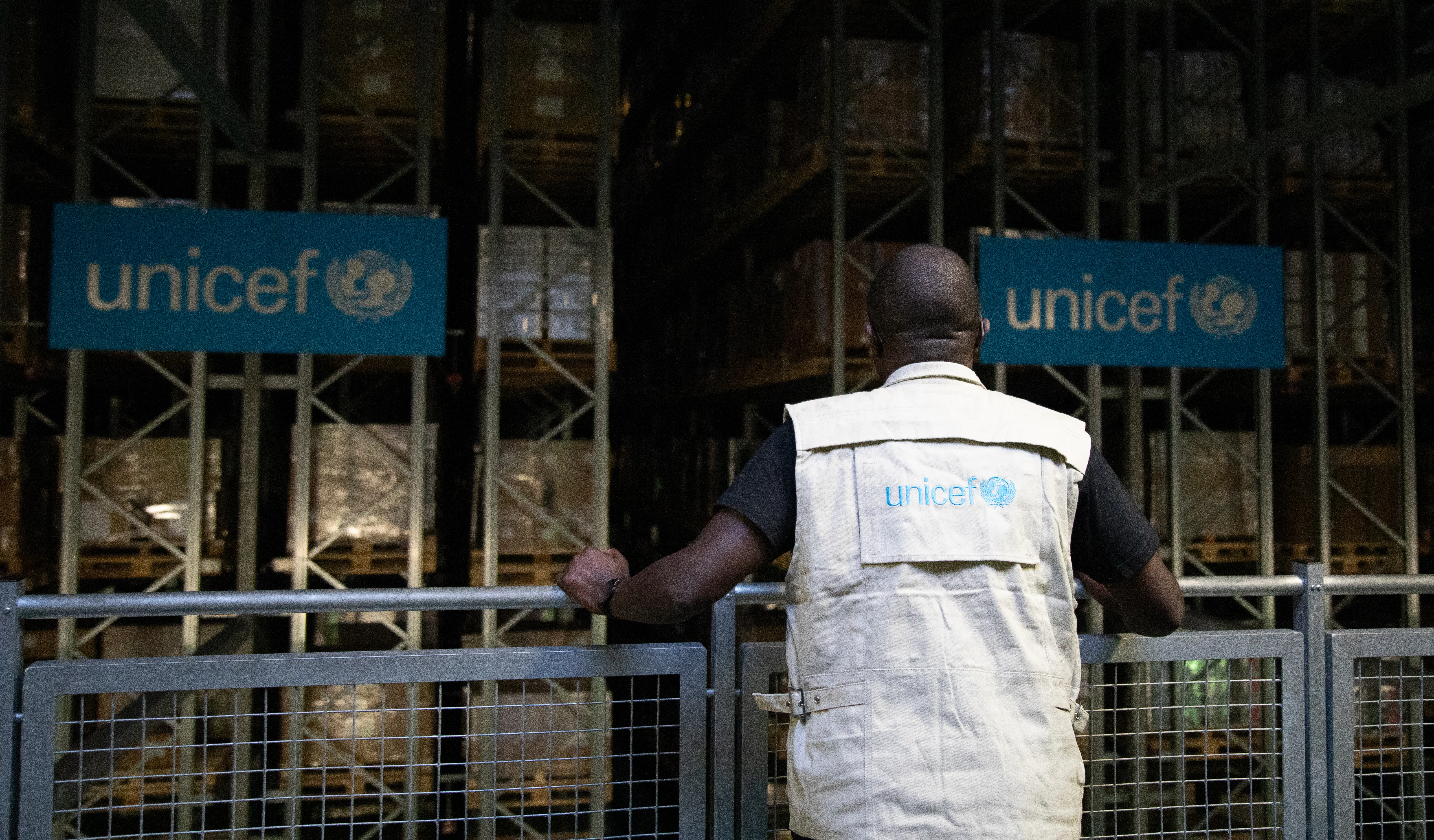 A man stands at a railing with his back to the camera, overlooking a warehouse staked with boxes. He is wearing a vest that says UNICEF.