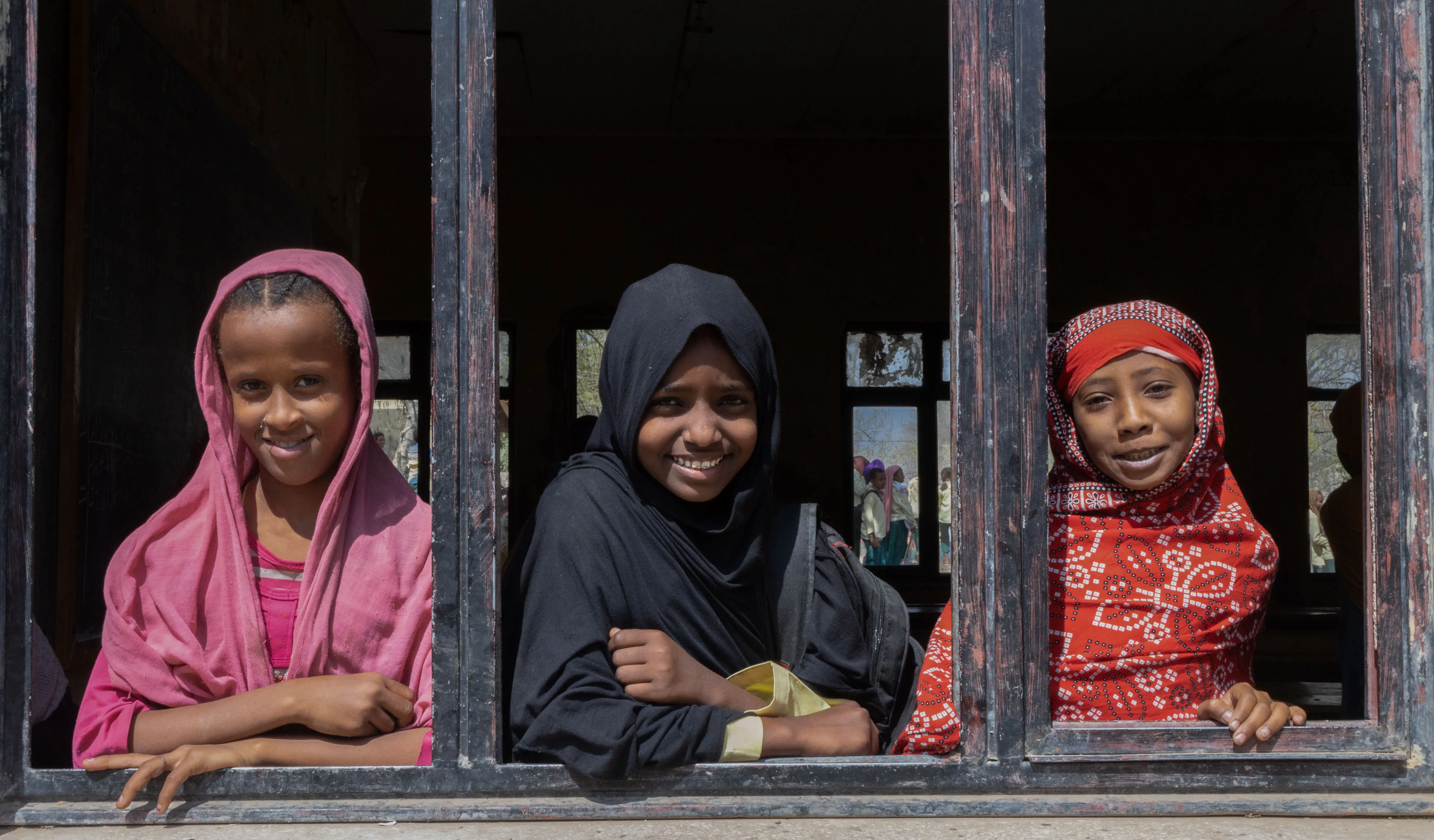 Three young girls with colourful head scarfs on, look out of a wooden window frame - all are smiling.  
