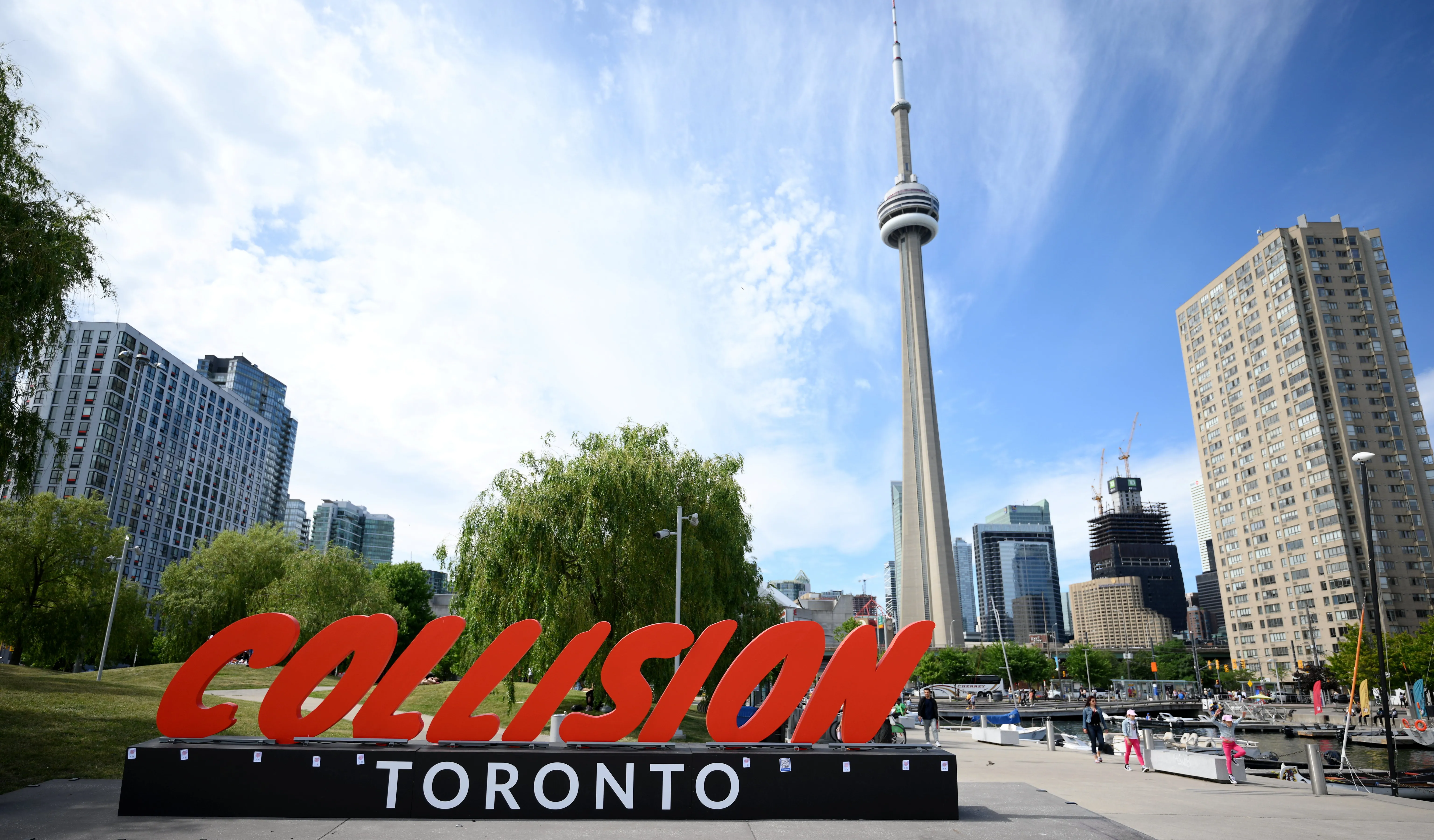 A red and black sign in the foreground reads 'Collision Toronto' in the background the CN tower and some buildings can be seen.  