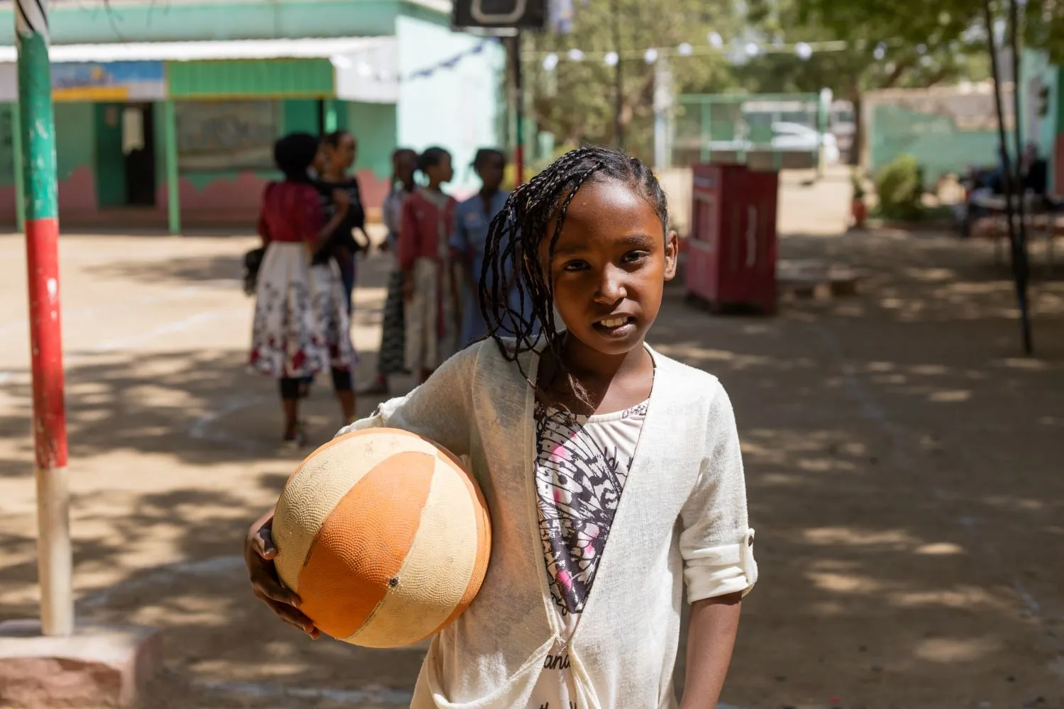 A child stands holding a basketball while smiling at the camera in an outdoor setting with other children standing in the background
