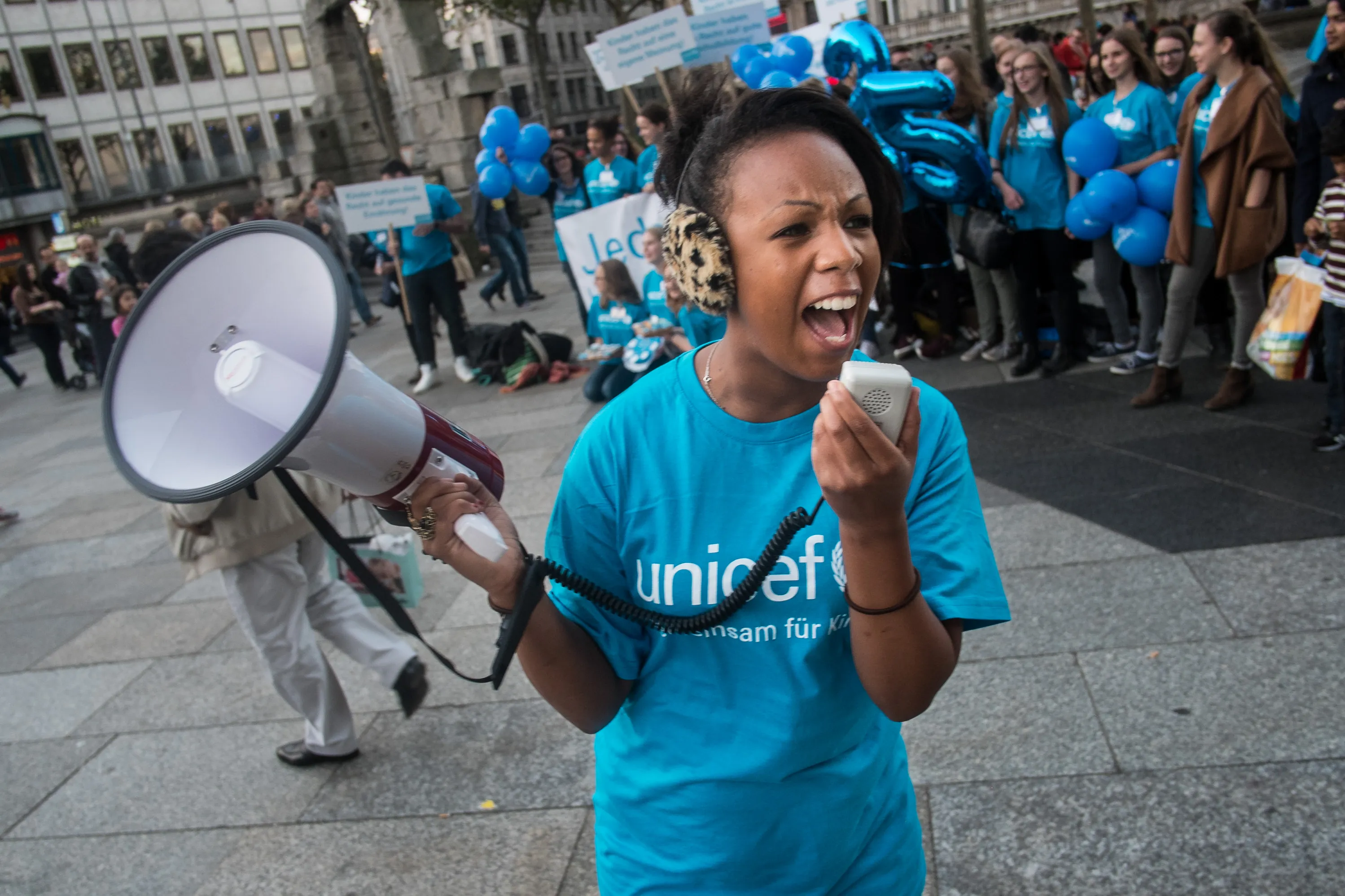 A girl wearing a blue UNCIEF tshirt speaks into a speakerphone, behind her others can be seen standing in a line also wearing UNICEF tshirts. 