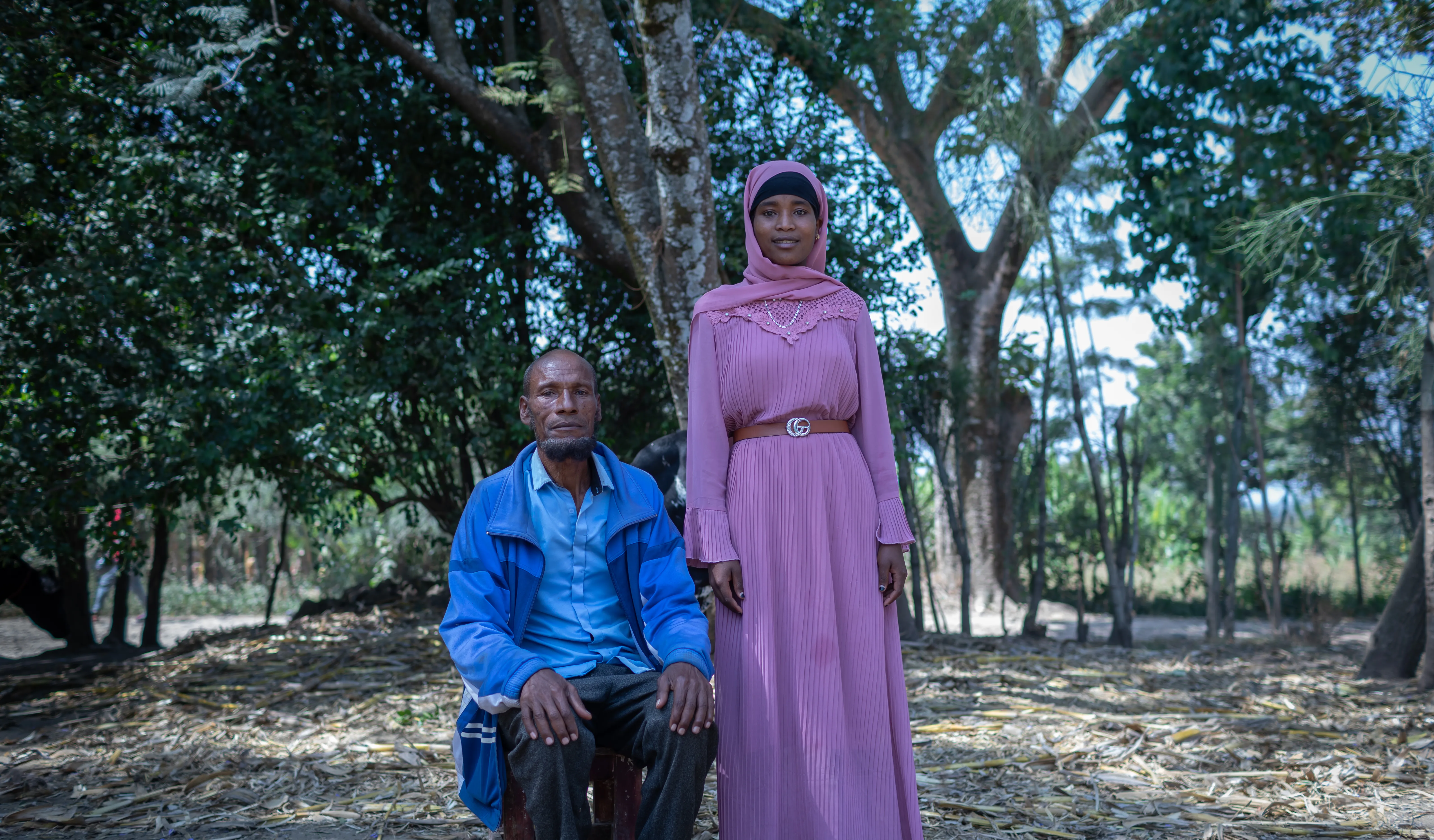 A man sits on a stool and his teenage daughter stands next to him. They are outdoors with many trees behind them.