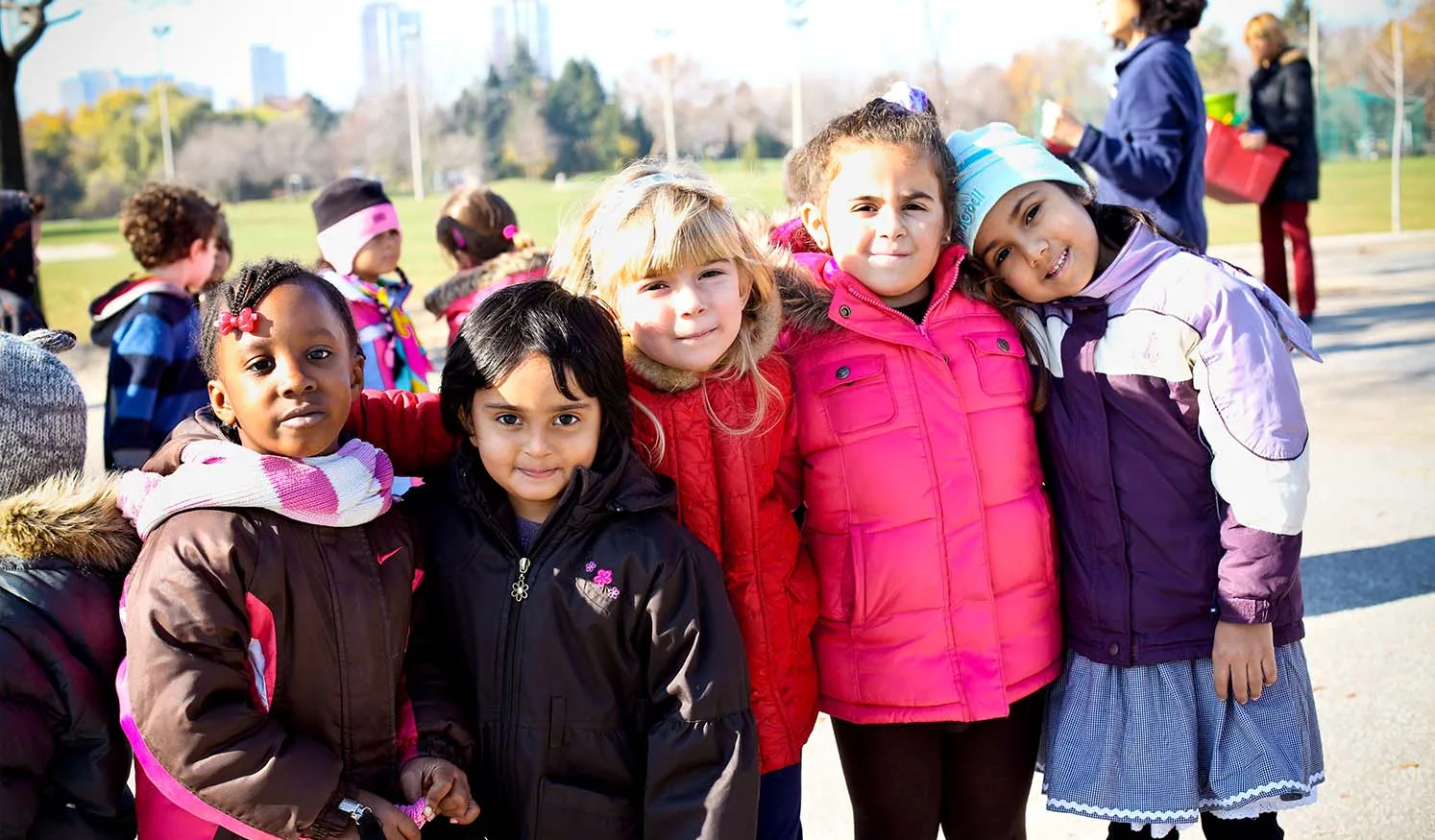 Des enfants aux origines diverses se tiennent côte à côte à l’extérieur par une journée fraîche et ensoleillée, en souriant à la caméra.