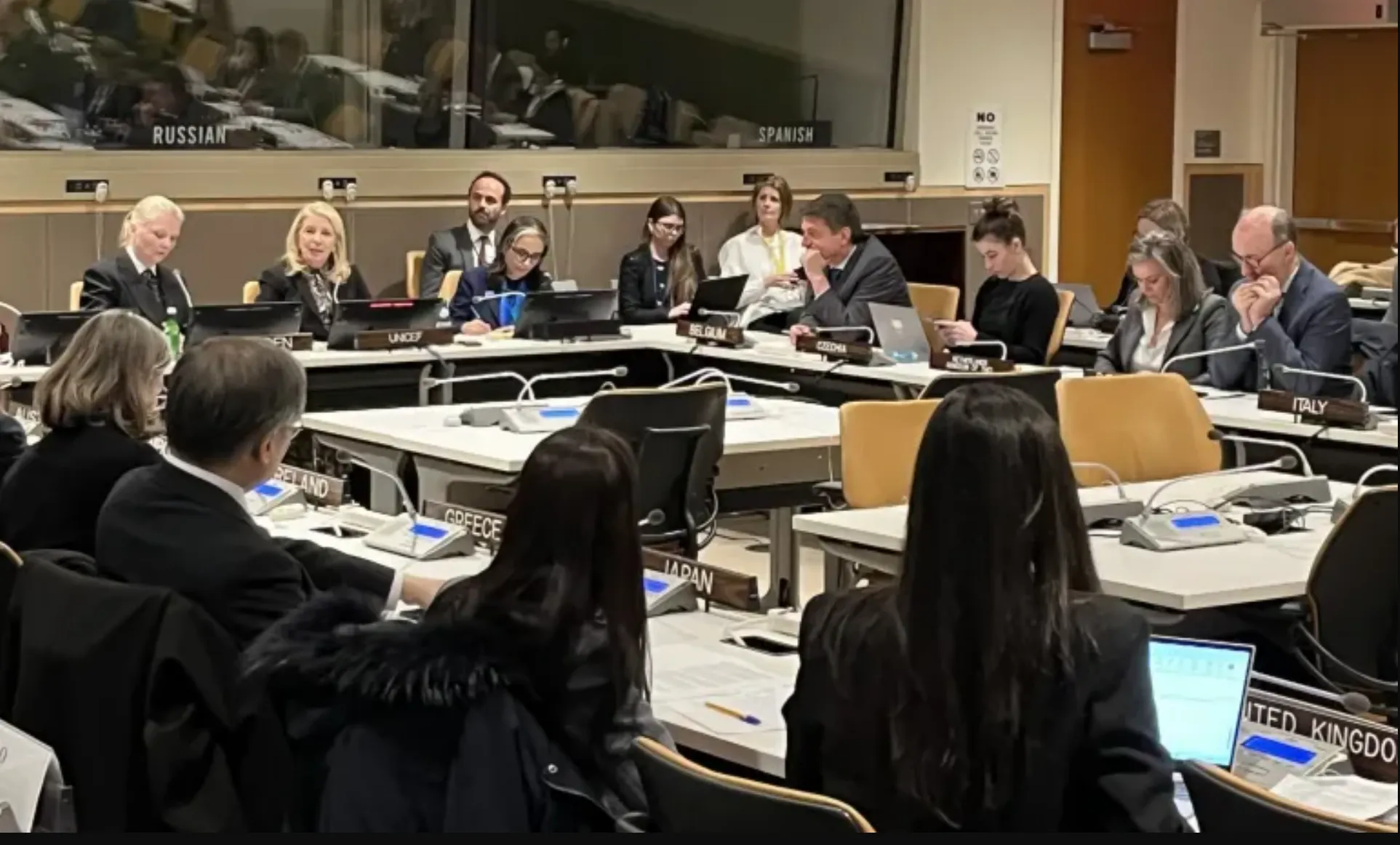 A group of delegates sit around a conference table with nameplates, engaged in discussion. The setting is formal, suggesting a diplomatic meeting.