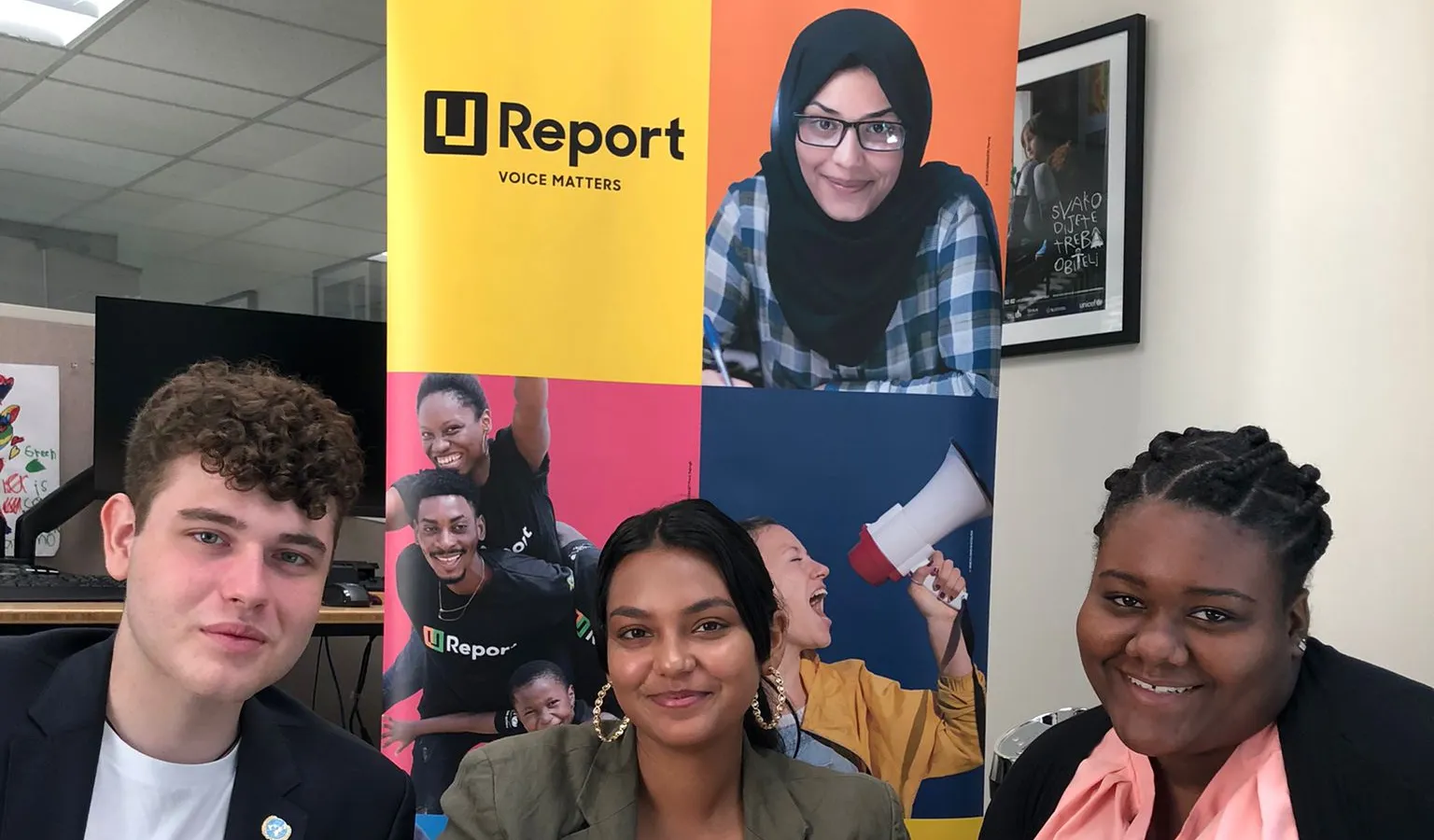 Three young people holding up cellphones in their hands and smiling. Behind them is a standee with a poster that shows the U-Report logo. 