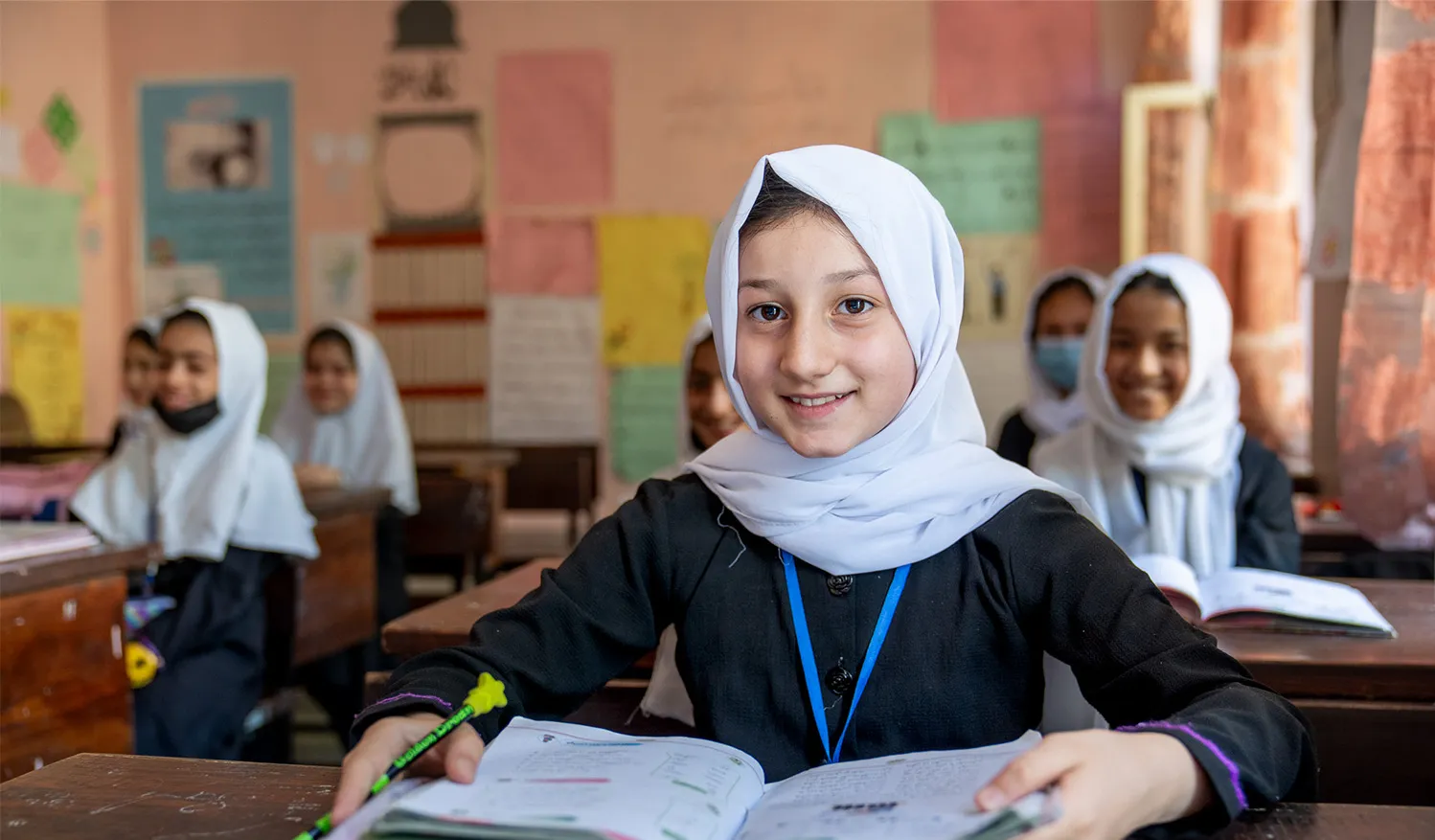 A girl with her textbook open smiles at the camera while sitting in a classroom in Kabul, Afghanistan.