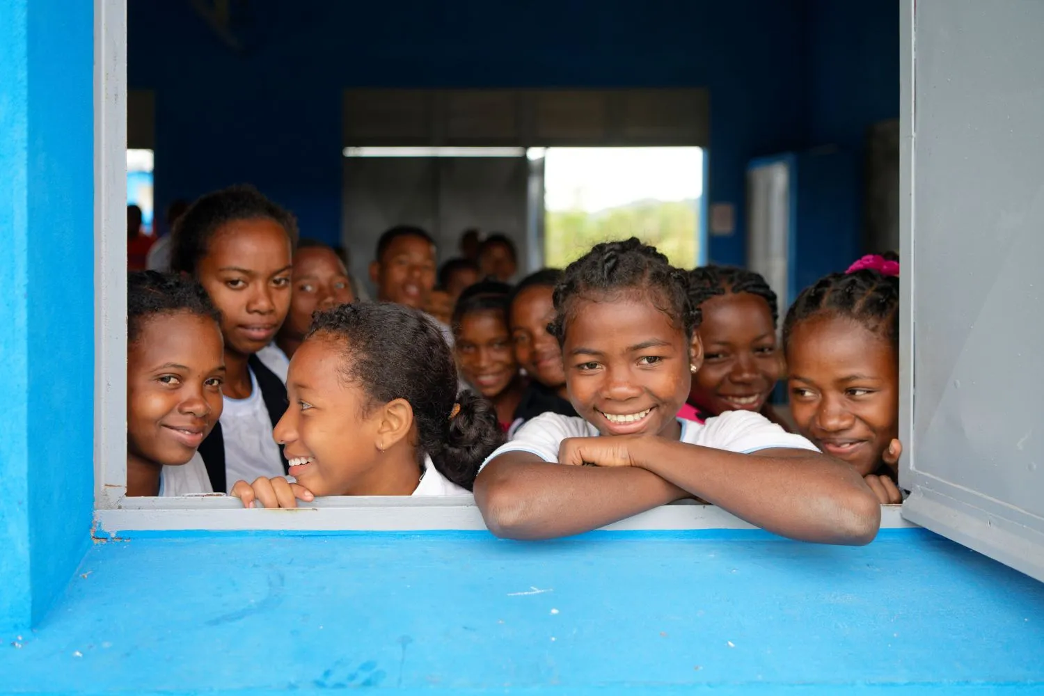 Un groupe de plus de dix enfants souriants regardent dehors depuis la fenêtre d’une salle de classe.