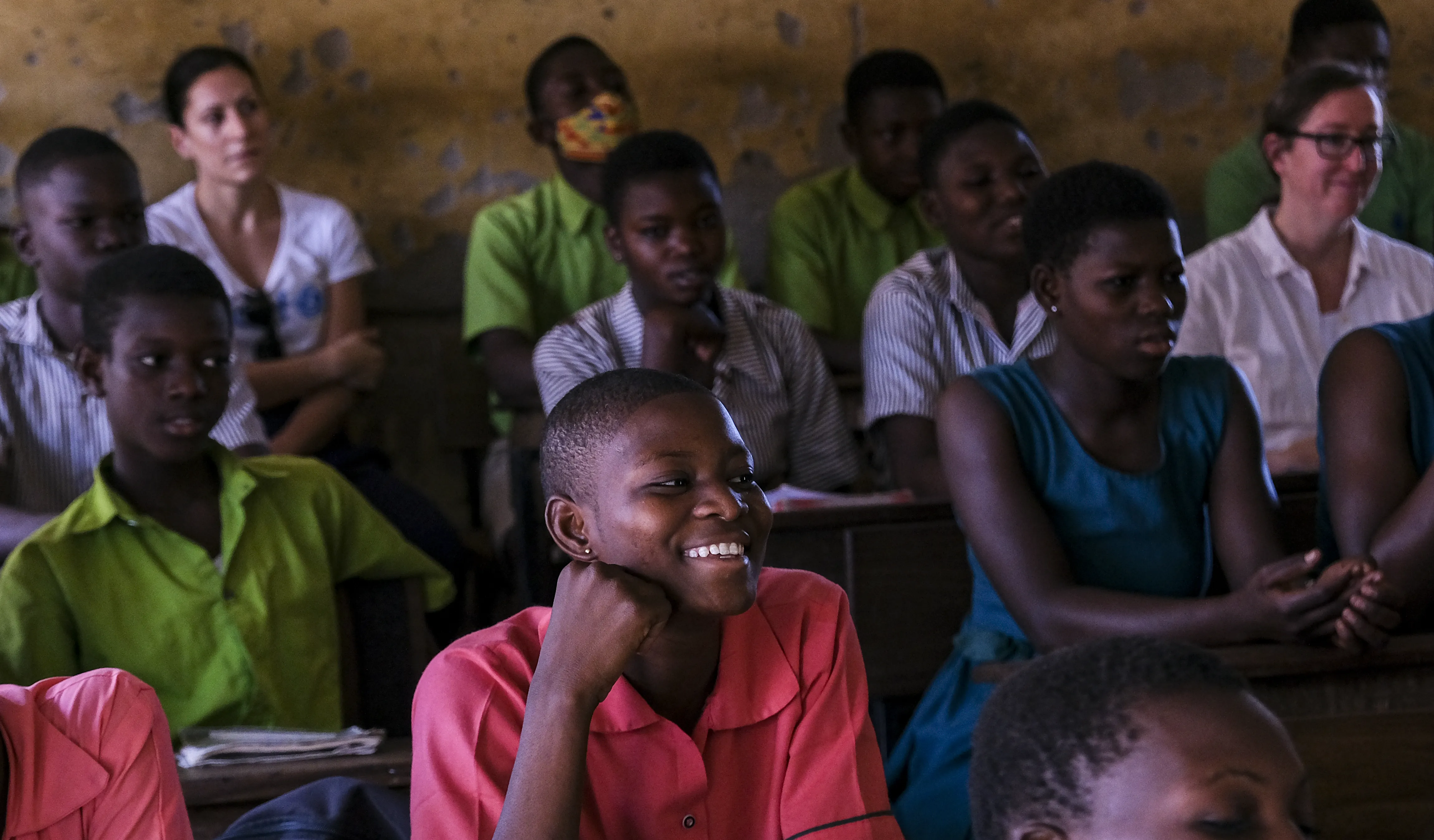 A classroom in Ghana with students sitting at desk. In the centre of the photo is a girl in a pink uniform smiling while looking beyond the camera. 