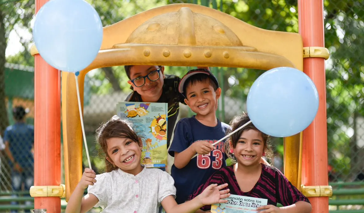 Four children stand together on a play structure and smile while holding balloons and reading materials.