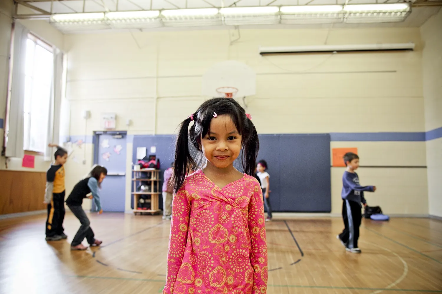 A young girl stands smiling at the camera on a basketball court, while her classmates play in the background. 
