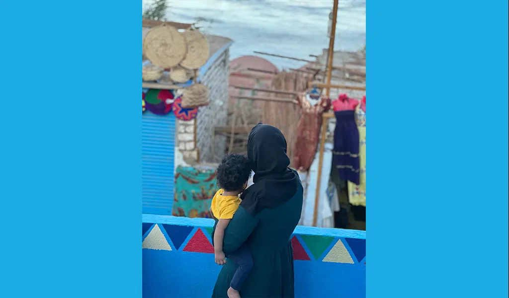 A woman looks out from a balcony, her back is towards the camera and she is holding a child in her arms. 