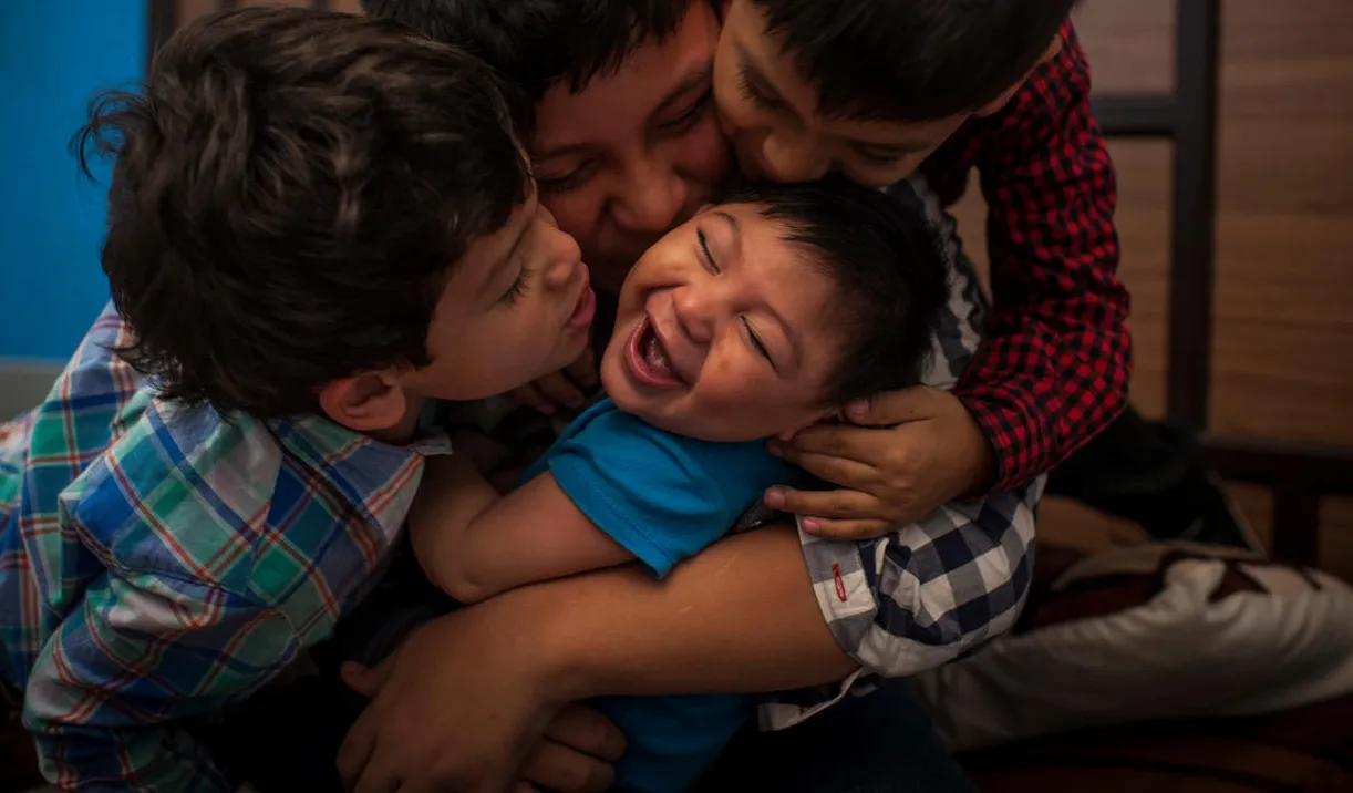 A boy laughs as his brothers shower him with kisses. Help UNICEF Support Right to a Childhood.
