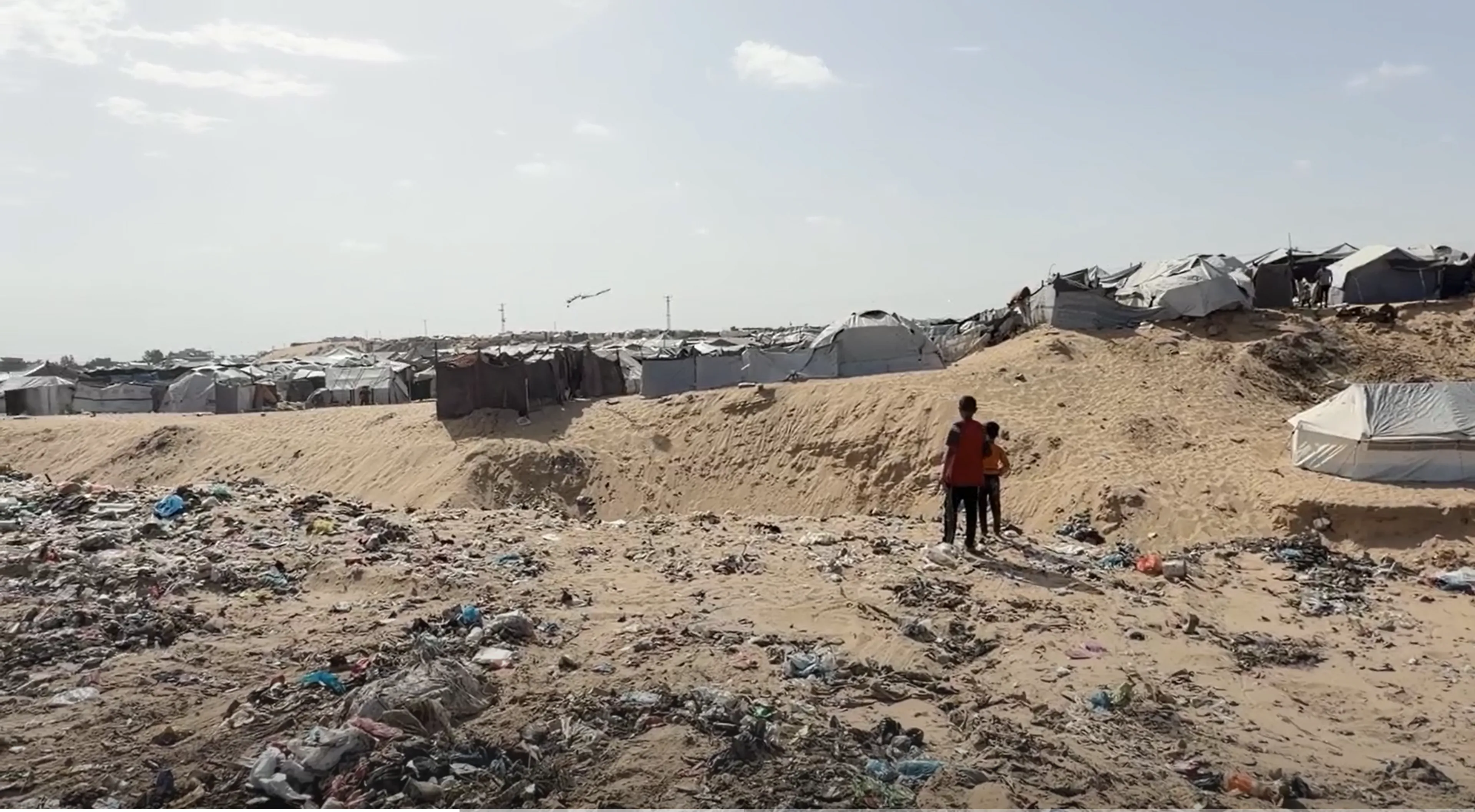 Two children stand in a sandy field with garbage around overlooking tents