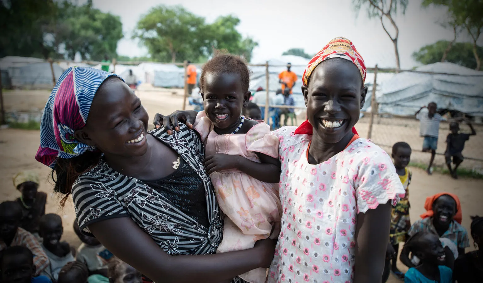 Two women smile, while looking at the camera, in between them is a child in a dress also smiling, the child is being held by the woman on the left. There are white tents behind them. This photo was taken in South Sudan in 2015. 