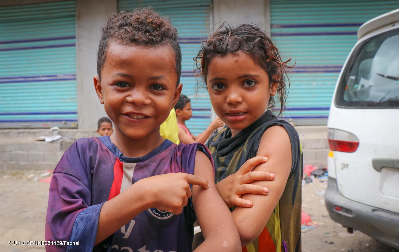 Two children on a street in Yemen, show off their arms with their sleeves rolled up, poking their arm to mimic a vaccine shot with their hands. 