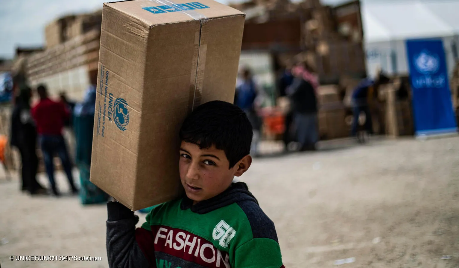 A child in a refugee camp carries a box of UNICEF supplies as he walks. This photo was taken in Syria in 2019. 
