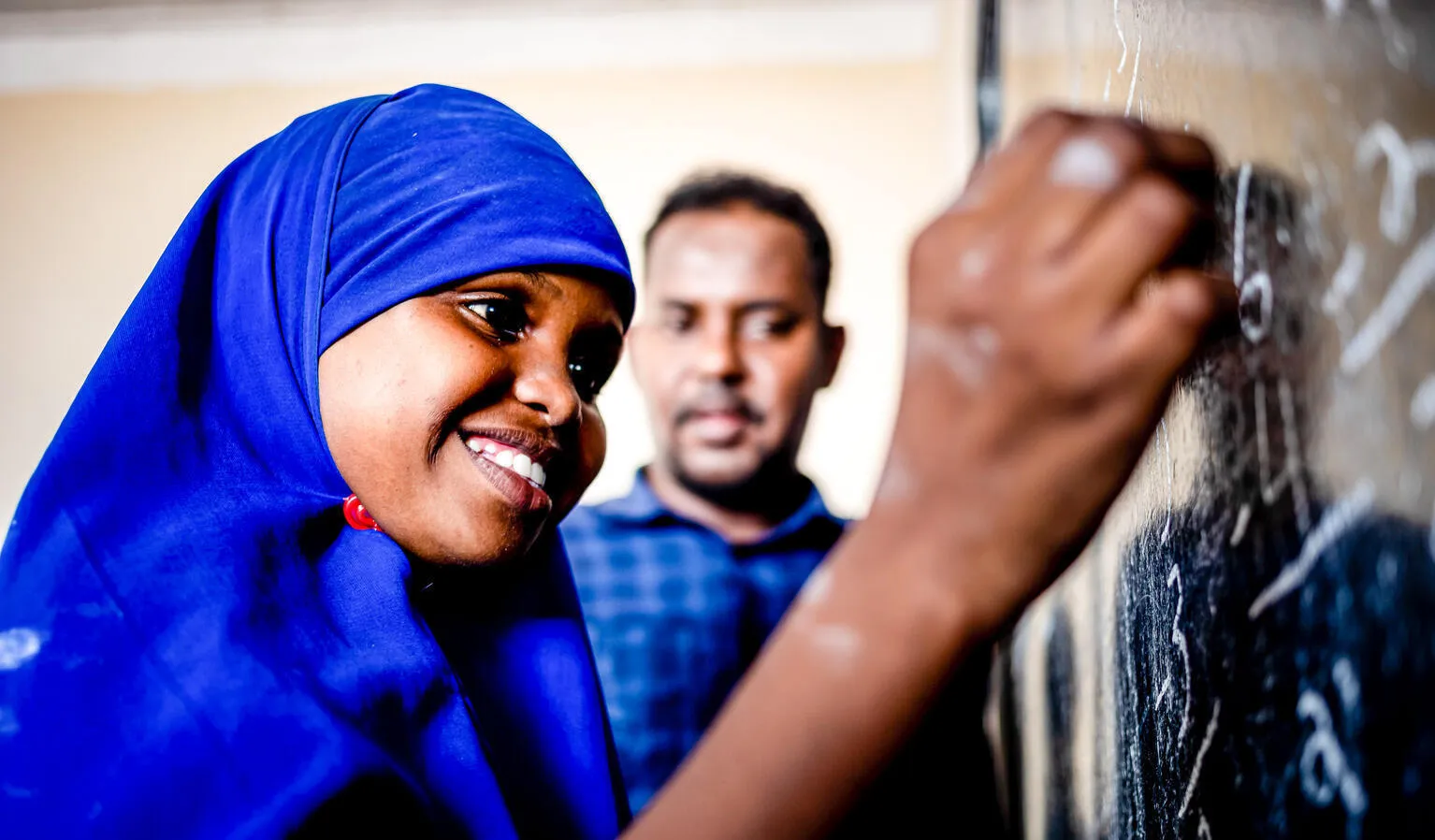 A girl wearing a blue uniform smiles as she writes on a chalk board as her teacher looks on in the background. This photo was taken in Somalia in 2021. 