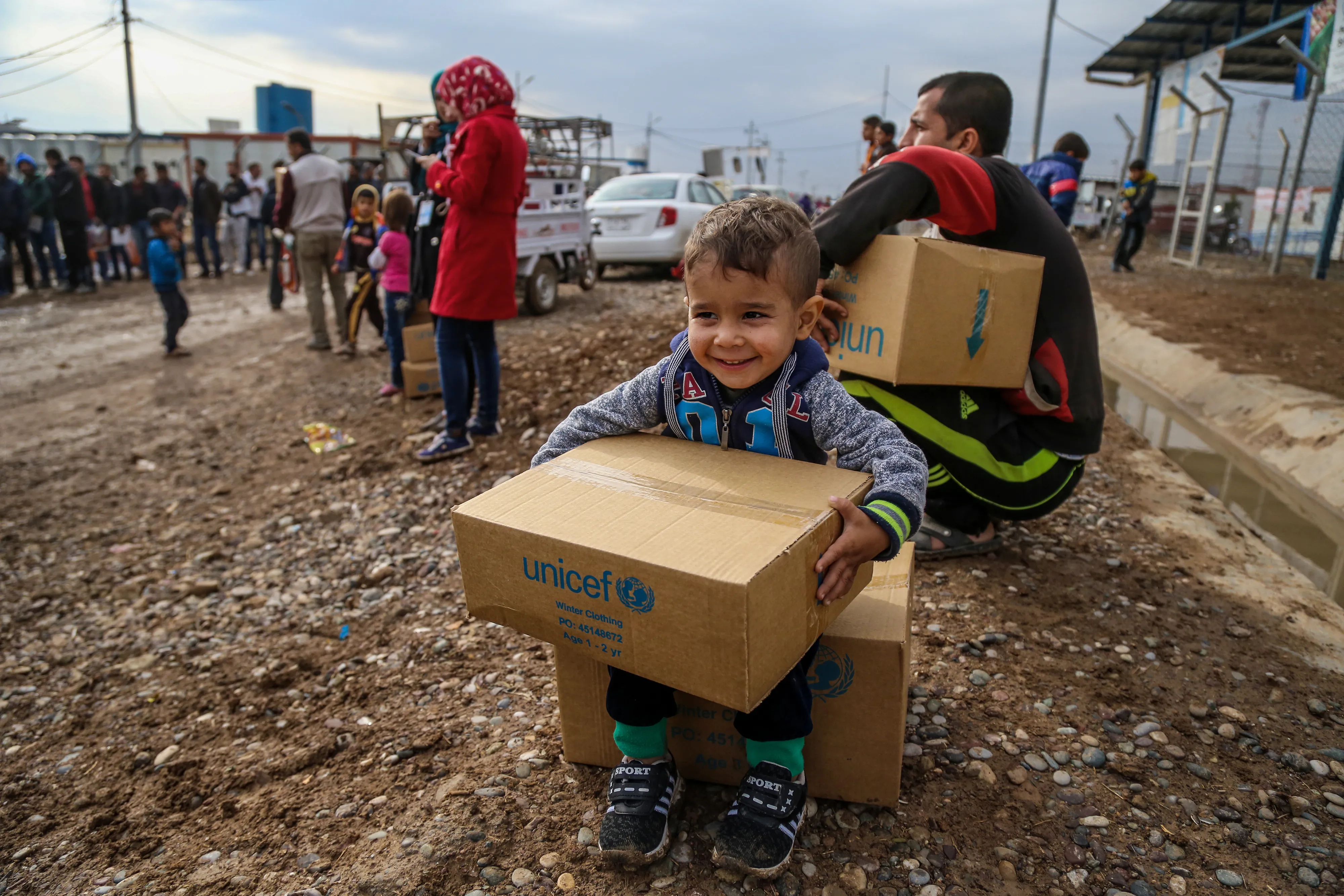 Smiling toddler sitting on a UNICEF supply box at a refugee camp distribution site.