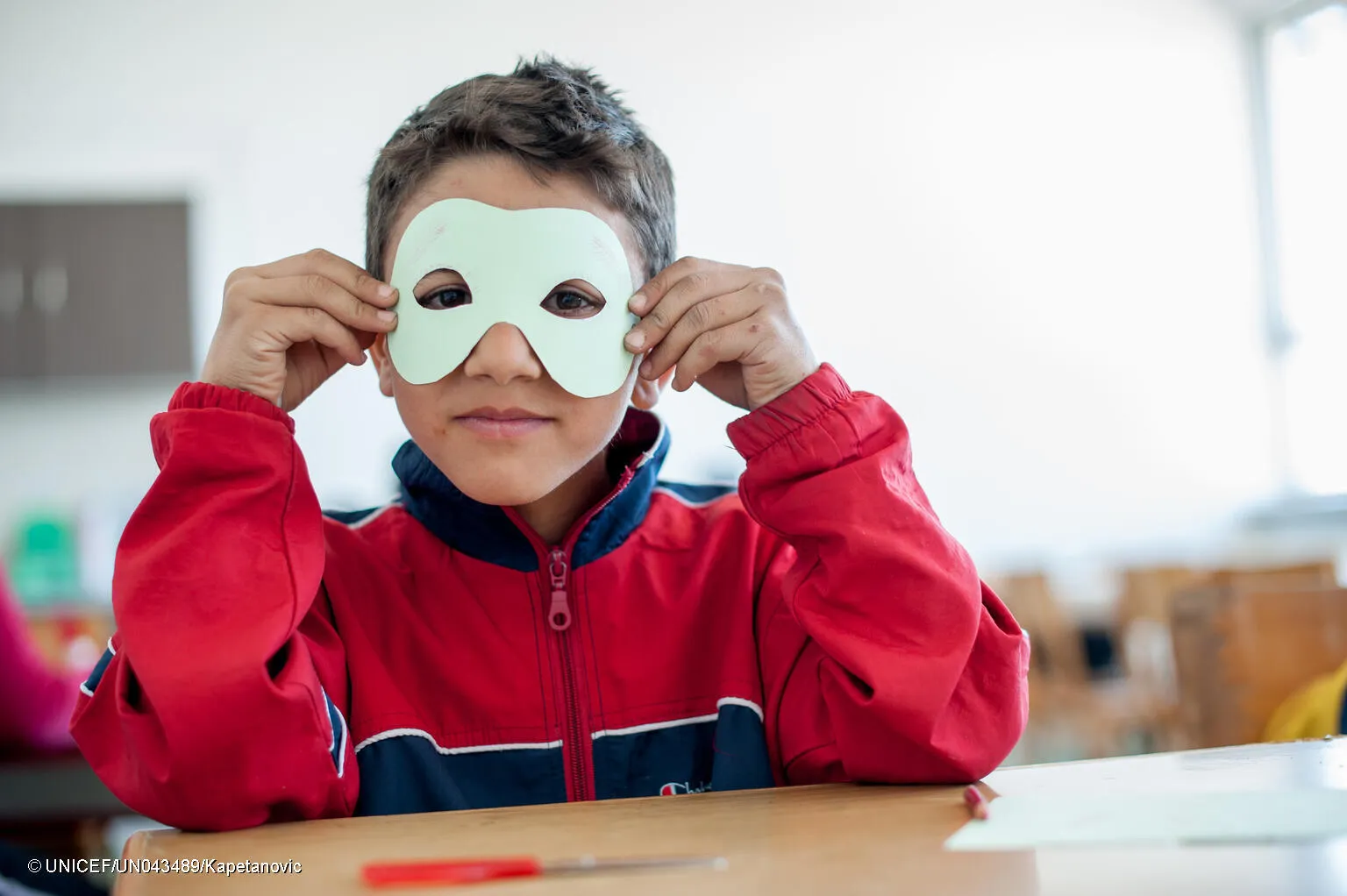 A child wearing a red jacket sits in a classroom desk. He is holding up a white paper mask with eyes cut out and holding it to his eyes. This photo was taken in Bosnia and Herzegovina in 2013. 