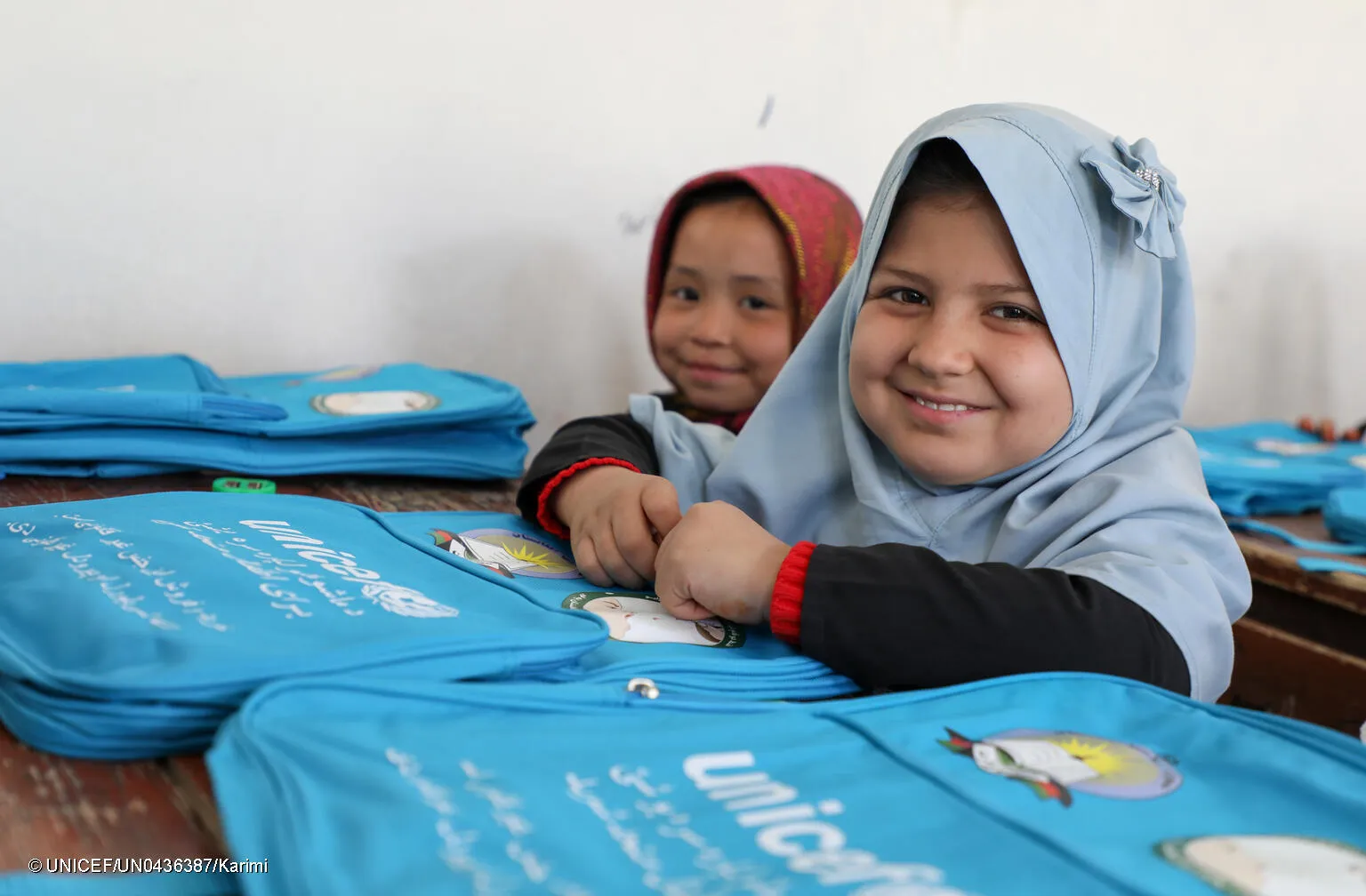 Two girls sitting at a desk with UNICEF bagpacks in front of them, both are looking at the camera and smiling. This photo was taken in Afghanistan in 2021. 