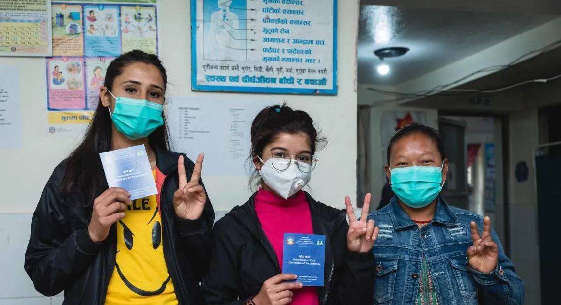 Three female health workers give the 'v for vaccinated' sign outside a clinic in Nepal.